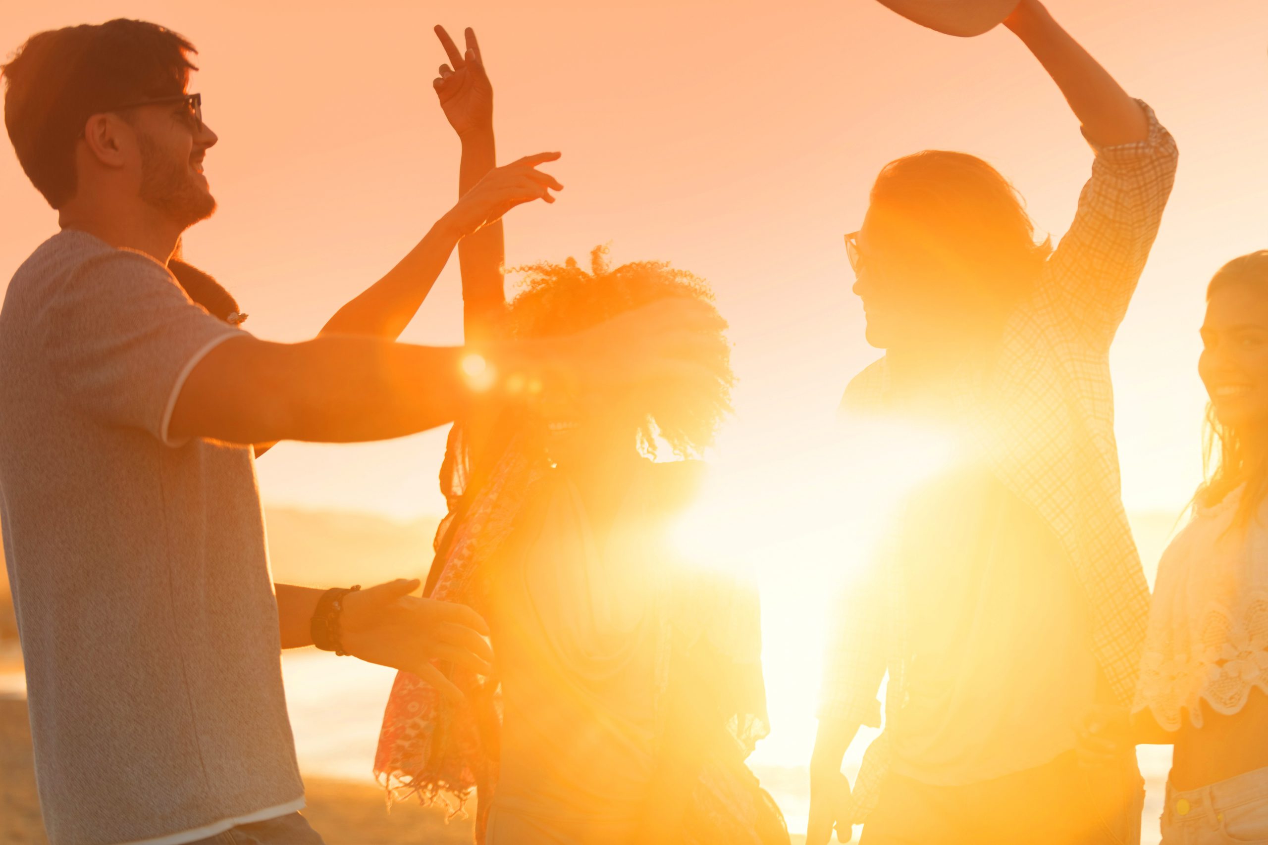 Group of friends dancing and celebrating on the beach at sunrise/sunset. They are very happy, smiling and laughing. Some have their arms raised. The sea and sun can be seen in the background. Defocussed with focus on background
